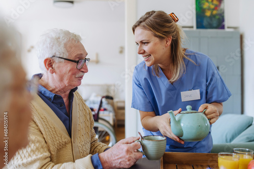 Happy healthcare worker holding teapot and talking to senior man at table photo