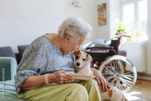 Happy senior woman holding plate of food and stroking dog at home photo