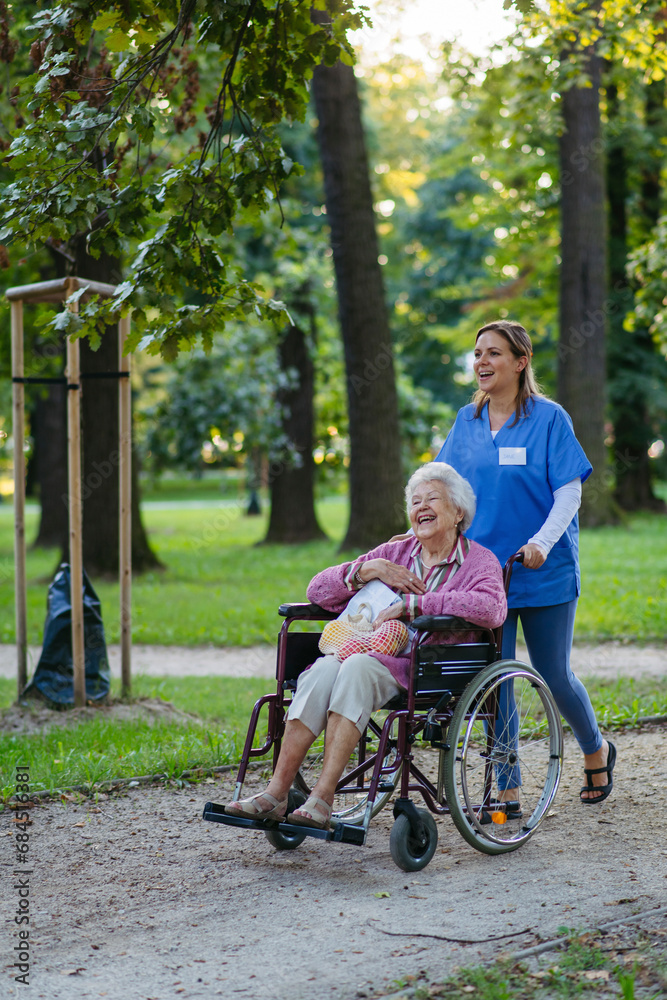 Happy healthcare worker walking with senior woman sitting in wheelchair at park Happy healthcare worker walking with senior woman sitting in wheelchair at park