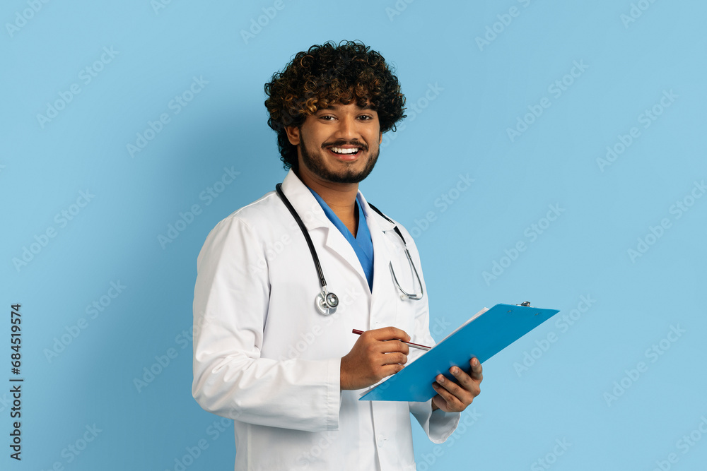 Positive indian man doctor in uniform holding clipboard and smiling Positive indian man doctor in uniform holding clipboard and smiling
