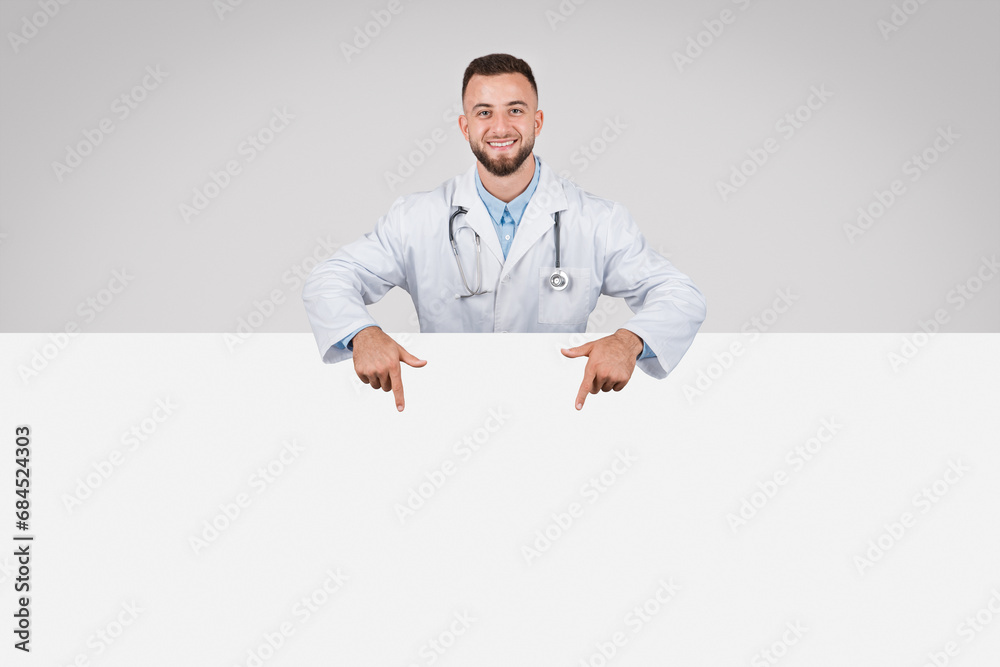 Doctor man pointing at empty white board below, grey background Doctor man pointing at empty white board below, grey background