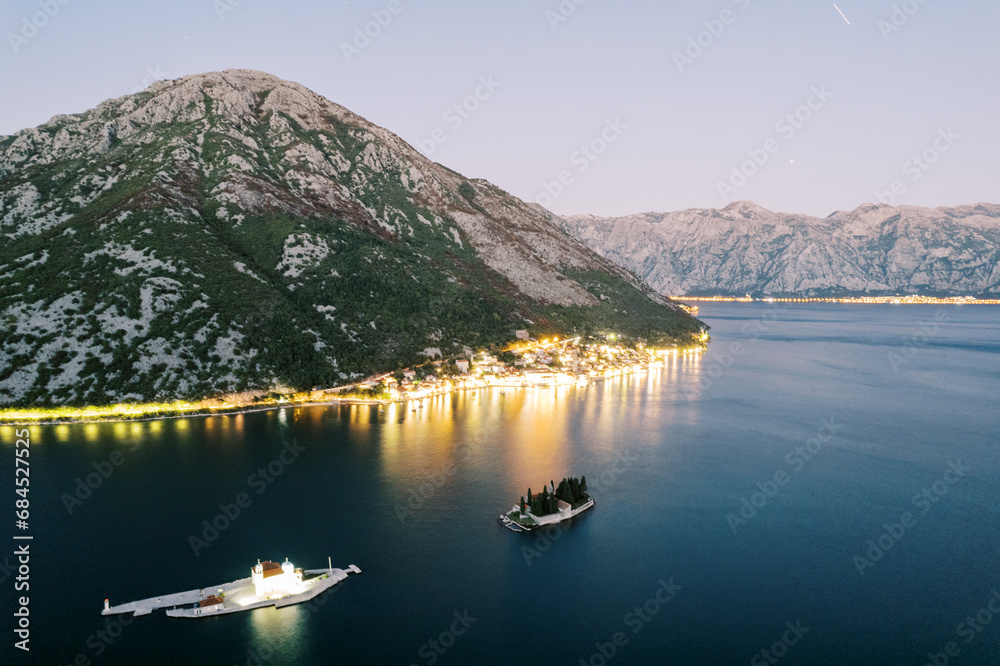 Illuminated Church of Our Lady on the Rocks on the island against the backdrop of illuminated Perast. Montenegro. Drone