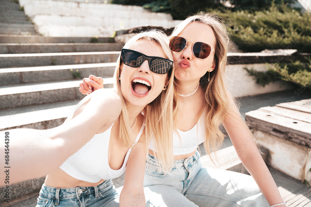 Two young beautiful smiling hipster female in trendy summer white top and jeans clothes. Carefree women posing in the street. Positive models having fun. Cheerful and happy. take selfie. In sunglasses