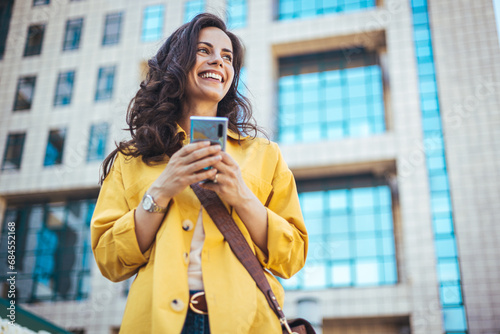 Beautiful happy young woman messaging on smartphone on the background of a city street on a sunny day. Business woman is looking away and smiling. 