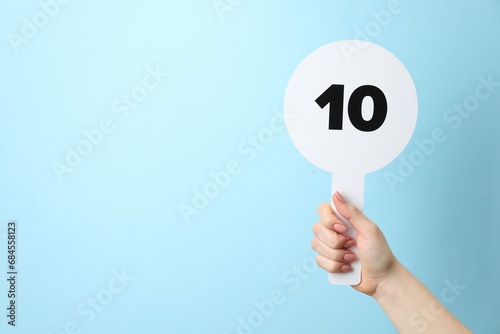 Woman holding auction paddle with number 10 on light blue background, closeup. Space for text