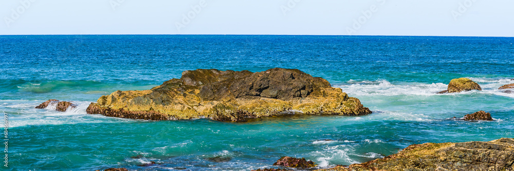 Volcanic rocks sticking out of the blue water at the shore, view from the beach of the sea landscape on a summer sunny day.