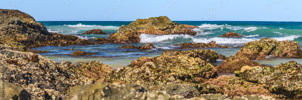 Australian coast with volcanic rocks at the shore, view from the beach to the horizon with blue water with waves on a summer sunny day.