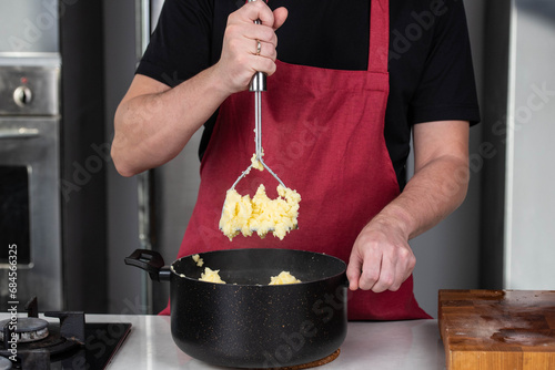 Man chef, donned in a red apron, skillfully mashes boiled potatoes in a pot, creating smooth, homemade mashed potato. The scene radiates culinary mastery and the joy of crafting a comforting dish. photo