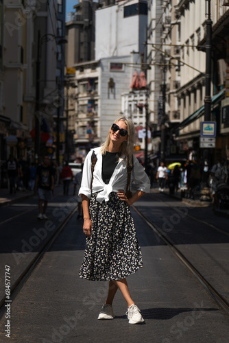 Beautiful blonde young woman in sunglasses poses on one of the main streets of Istanbul on a sunny day. Stylish female tourist with a backpack in Turkey. Vertical photo