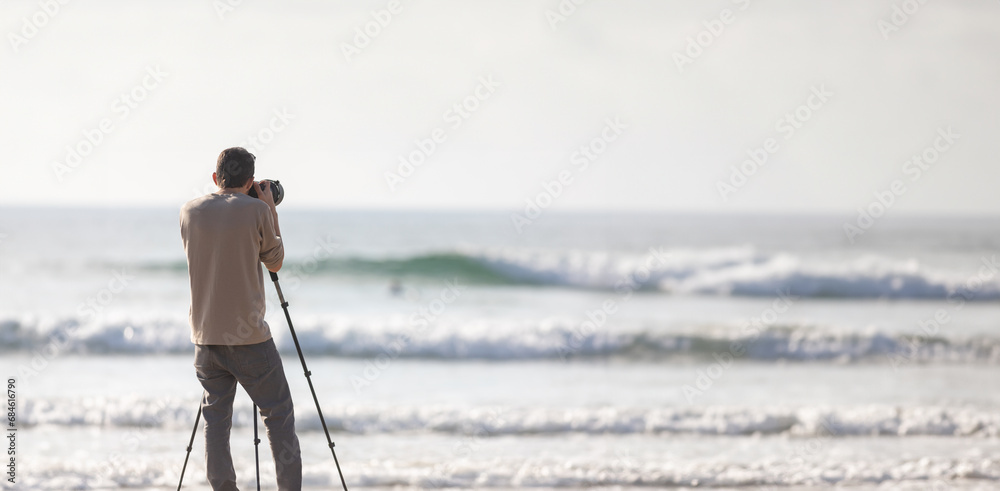 Young handsome photographer with a tripod and a camera on the beach - obrazy, fototapety, plakaty 