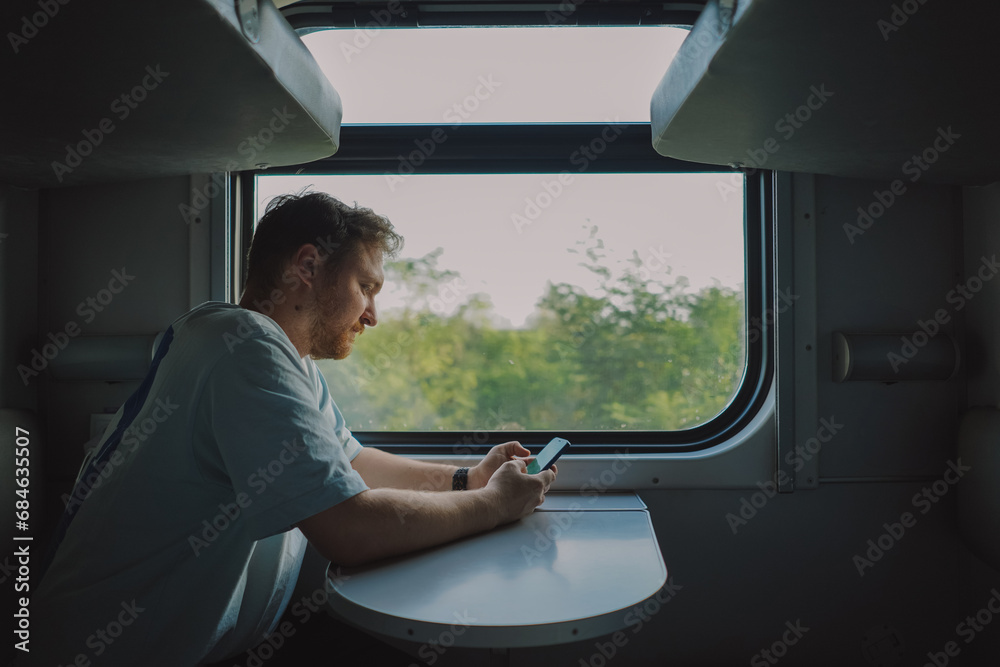 Fototapeta premium A man with a beard and mustache in a blue t-shirt is using a smartphone while traveling by Railway train, sitting in the train and looking out the window.