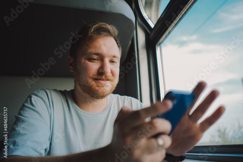A man with a beard and mustache in a blue t-shirt is using a smartphone while traveling by Railway train, sitting in the train and looking out the window. © Анастасія Стягайло