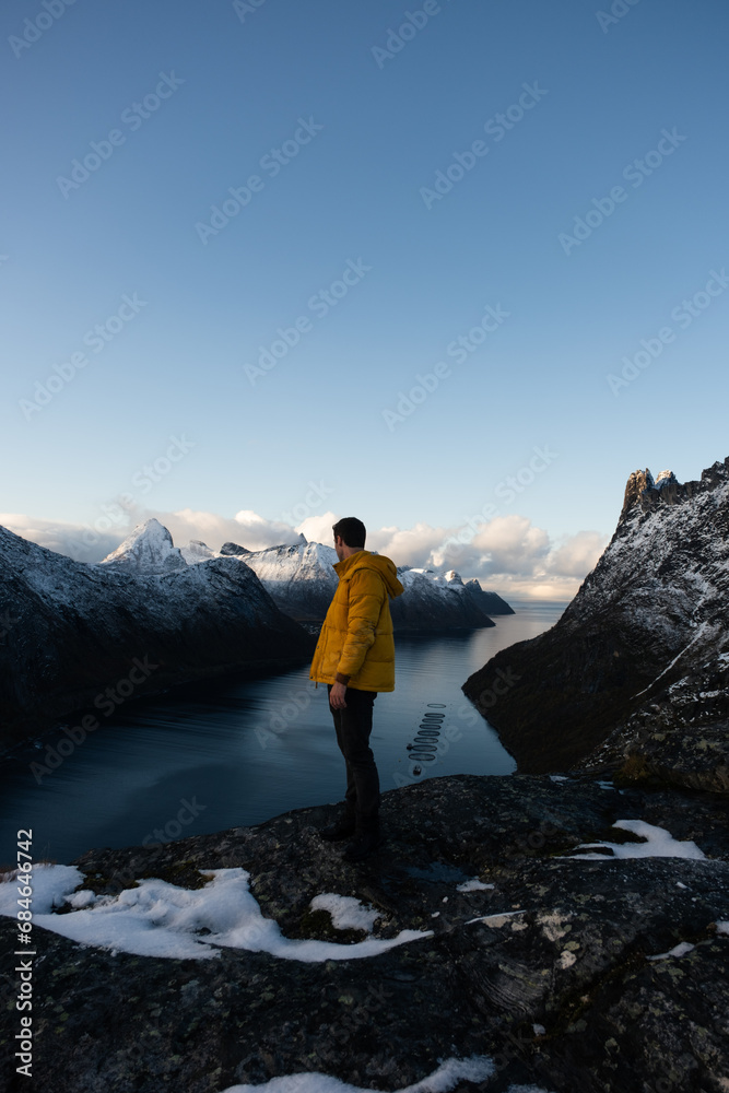 Man in yellow coat standing on mountain overlooking Norweigan Fjord.  Shot on a Mirrorless camera in the fall near Segla, Senja, Norway in the Arctic Circle.  Travel Influencer style photo.