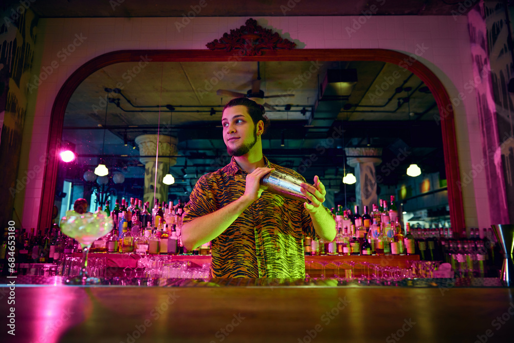Photo with selective focus of young guy shaking cocktail shaker making sweet tasty cocktails at bar with modern design and neon illumination. Photo with selective focus of young guy shaking cocktail shaker making sweet tasty cocktails at bar with modern design and neon illumination.