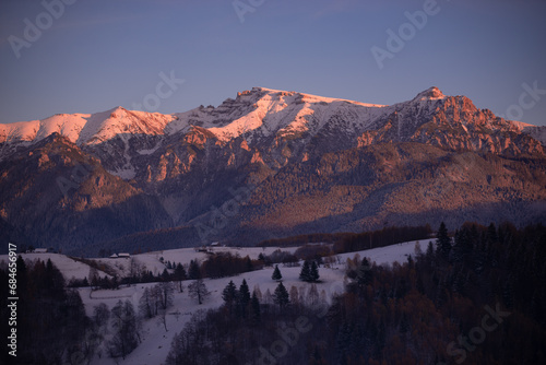 Fairy tale landscape with a rocky mountain full of snow at sunset. A village of traditional Romanian old houses spread over a valley.