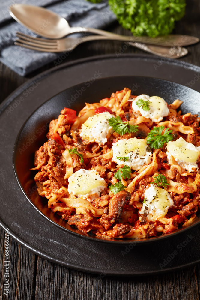 Lasagnette with beef ragu, mushrooms and ricotta cheese in black bowl on dark wooden table with fork and spoon at background, italian recipe, vertical view