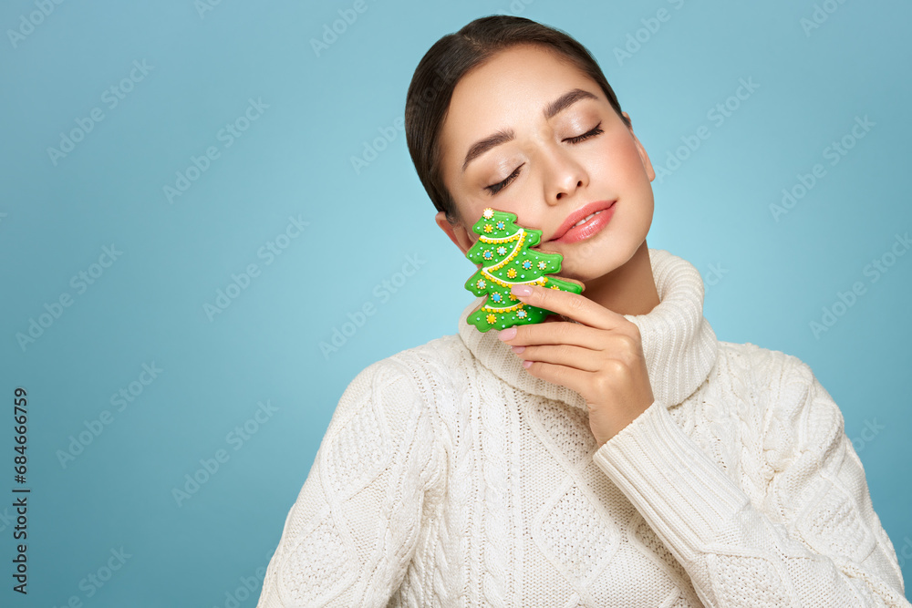 Woman in a white knitted sweater with cookie Christmas tree isolated on blue background. Christmas or New Year festivities. Holiday and Party Woman in a white knitted sweater with cookie Christmas tree isolated on blue background. Christmas or New Year festivities. Holiday and Party
