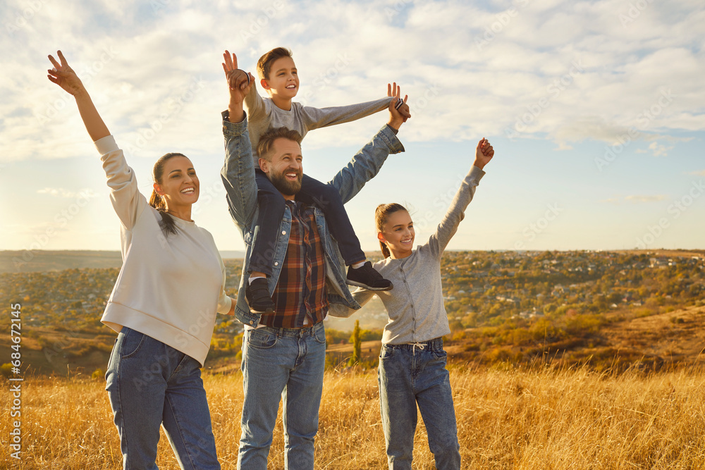 Portrait of happy joyful family standing in the field with two kids and looking into the distance with hands up enjoying beautiful nature together. Young parents with children walking outdoors. Portrait of happy joyful family standing in the field with two kids and looking into the distance with hands up enjoying beautiful nature together. Young parents with children walking outdoors.