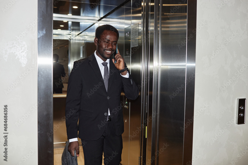 Medium full shot of smiling Black man wearing elegant business suit talking on smartphone while leaving office elevator, copy space Medium full shot of smiling Black man wearing elegant business suit talking on smartphone while leaving office elevator, copy space