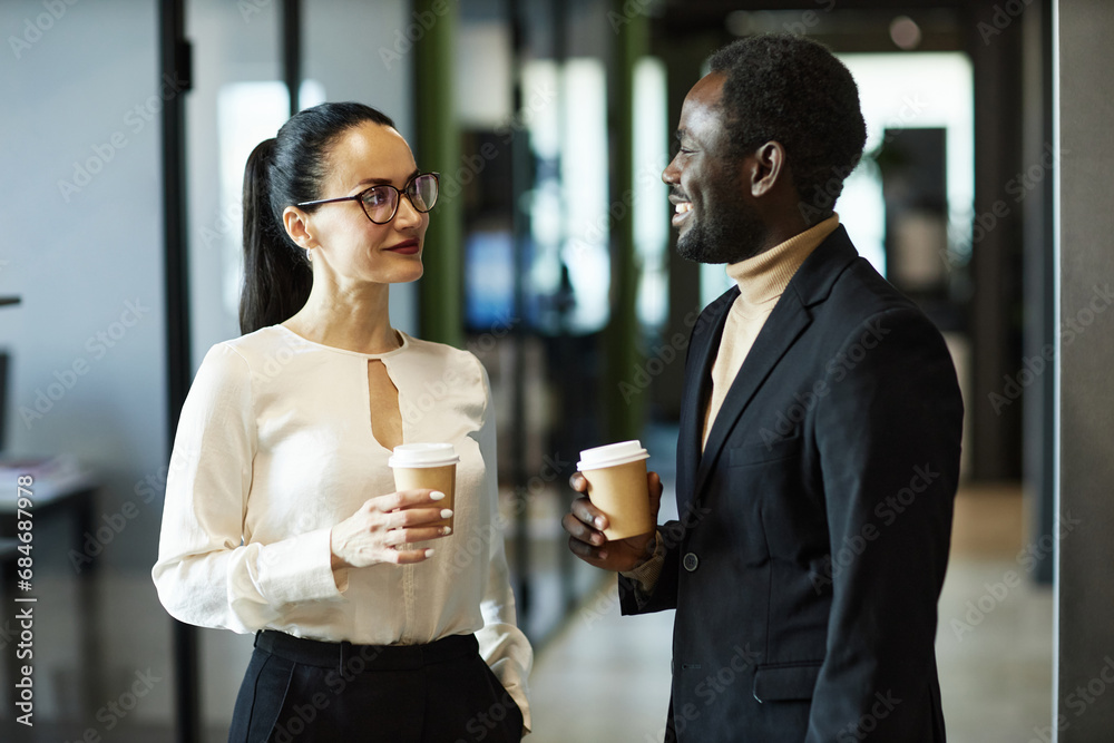 Waist up shot of Caucasian business lady and African American Businessman face to face having conversation drinking coffee standing in office hall Waist up shot of Caucasian business lady and African American Businessman face to face having conversation drinking coffee standing in office hall