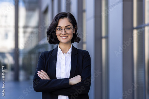 Close-up portrait of a young businesswoman standing outside an office building with her arms crossed over her chest. Confidently and smilingly looking at the camera photo