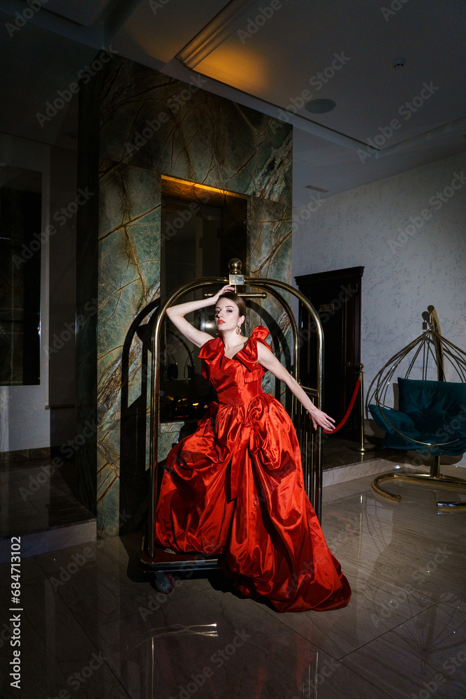 Pretty woman in a luxurious red dress posing in a luggage trolley.Fashion shooting concept Pretty woman in a luxurious red dress posing in a luggage trolley.Fashion shooting concept