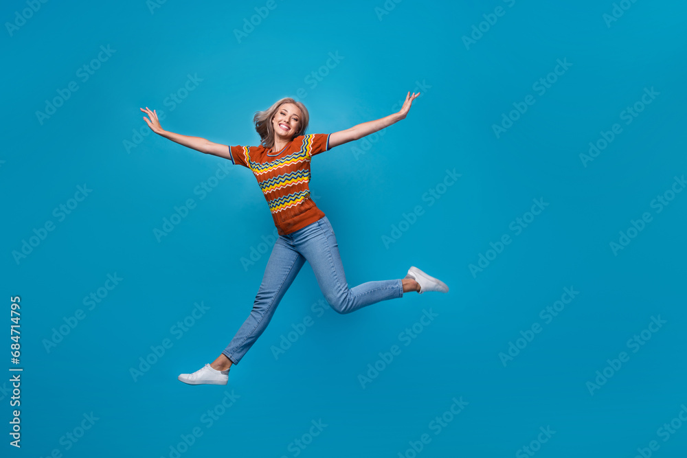 Full length photo of positive excited girl dressed print shirt jumping high arms sides empty space isolated blue color background