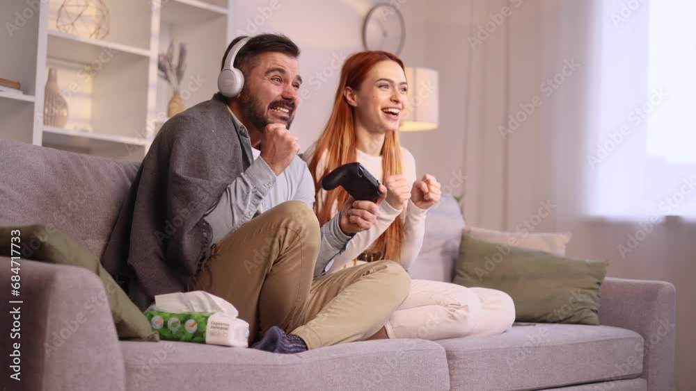 Excited handsome man sitting on the couch and playing in video games on digital console while his girlfriend wife supports and encourages boyfriend husband at home together Happy couple family concept Excited handsome man sitting on the couch and playing in video games on digital console while his girlfriend wife supports and encourages boyfriend husband at home together Happy couple family concept