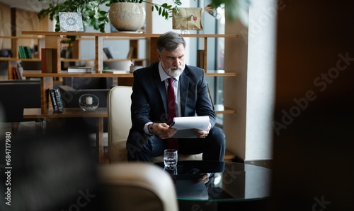 Mature businessman in formal suit concentrating on reading the report, sitting in the lobby and preparing for the meeting. © opolja