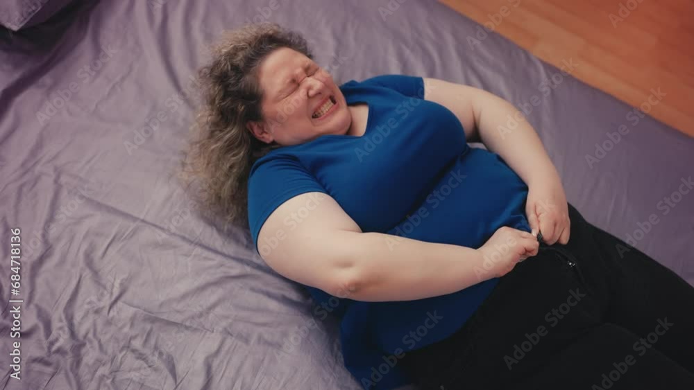 Top view of large woman lying on bed, unable to button tight jeans on big belly