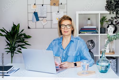 Portrait of 50 s confident mature businesswoman  middle-aged experienced senior female professional using phone  working on laptop in cozy office. Female CEO  female entrepreneur manage business.