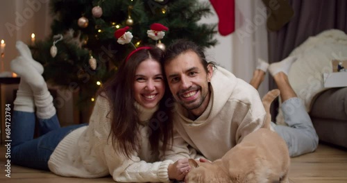 Portrait of a happy couple, a guy and a brunette girl in white sweaters lie on the floor near their cream-colored cat near the Christmas Tree in an apartment that is decorated in the spirit of photo