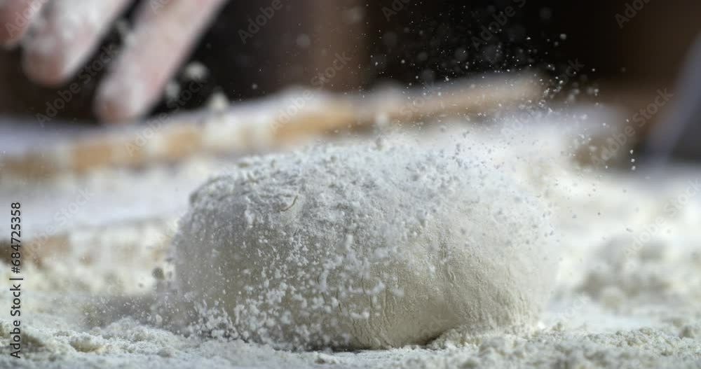 Super slow motion close up of professional artisan baker chef sprinkles flour on raw loaf of dough while making homemade bread, pasta or pizza on rustic wooden table in traditional bakery kitchen.