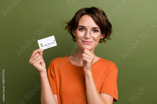Photo of clever positive girl dressed orange t-shirt in glasses holding credit card arm on chin isolated on khaki color background photo