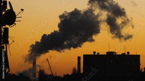 CHP chimney with a huge column of smoke and silhouette of a residential apartment block. Beautiful orange colours of predawn. Global energy crisis. Cityscape of a cold morning.