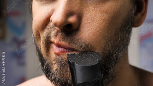 A man trims his beard with a trimmer. Close up. An electric razor trimming a beard. Black beard with grey hairs. Beard care at home.