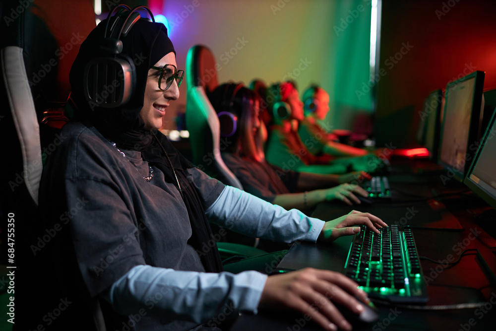 Side view portrait of smiling Muslim woman playing video games in cybersports club with neon lighting, copy space