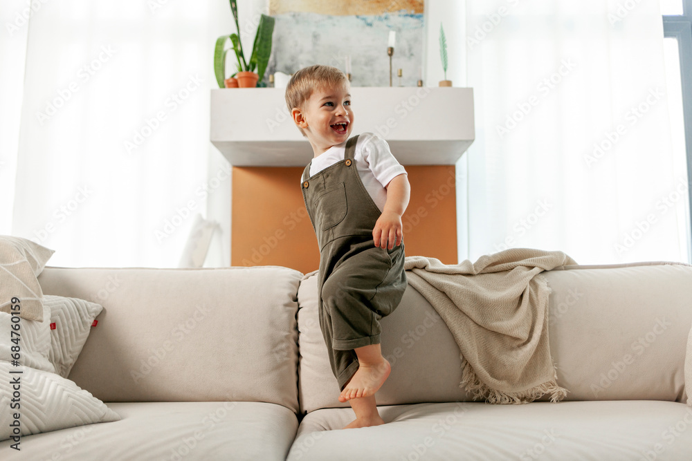 little boy 2 years old stands on soft comfortable sofa at home and laughs, happy child smiles and looks away little boy 2 years old stands on soft comfortable sofa at home and laughs, happy child smiles and looks away