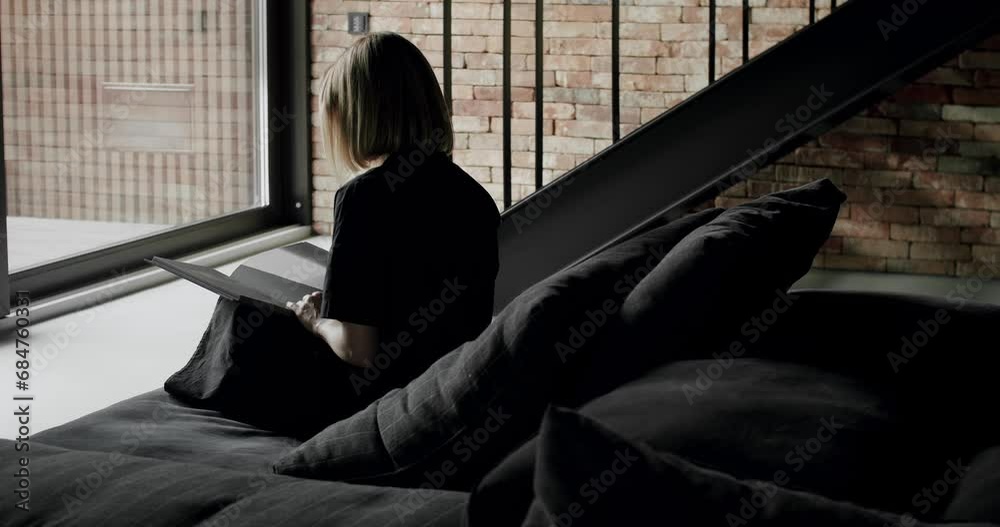 Back view on young woman sitting on comfortable sofa with cushion and reading book while resting at home. Female watching catalog on modern couch in apartment with loft interior Back view on young woman sitting on comfortable sofa with cushion and reading book while resting at home. Female watching catalog on modern couch in apartment with loft interior