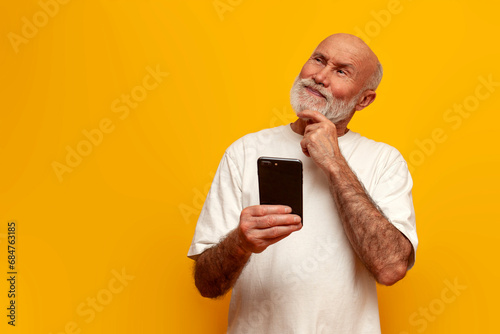 pensive old bald grandfather in white t-shirt uses smartphone and thinks on yellow isolated background