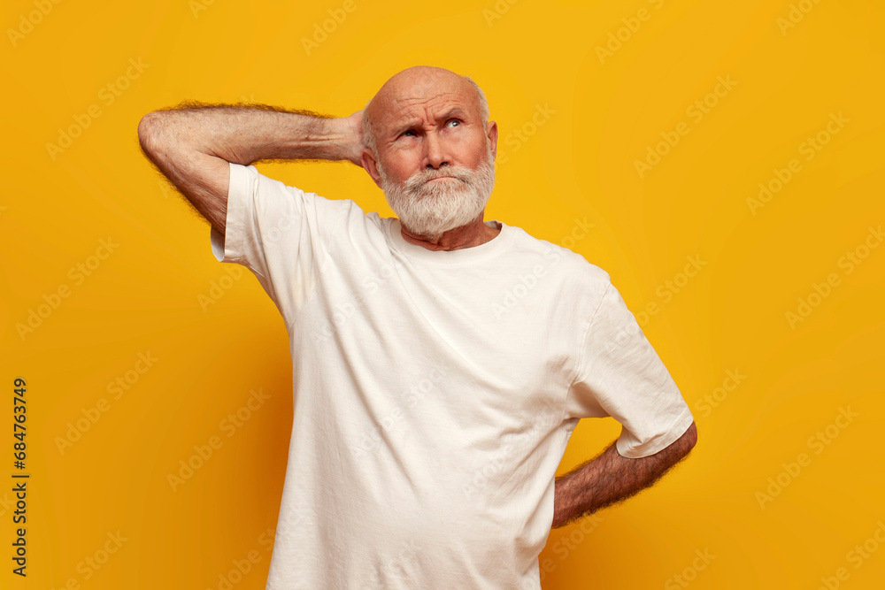 puzzled old bald grandfather in white t-shirt thinks remembers and scratches his head on yellow isolated background puzzled old bald grandfather in white t-shirt thinks remembers and scratches his head on yellow isolated background
