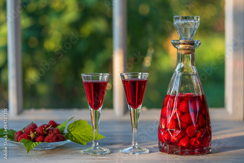 Homemade red raspberry brandy in two wine glasses and in a glass bottle on a wooden windowsill near summer garden, closeup