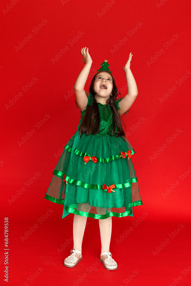 A girl in a festive carnival costume of little Christmas tree pointing at an empty space on a red background. Winter holidays concept. Sale and advertising A girl in a festive carnival costume of little Christmas tree pointing at an empty space on a red background. Winter holidays concept. Sale and advertising