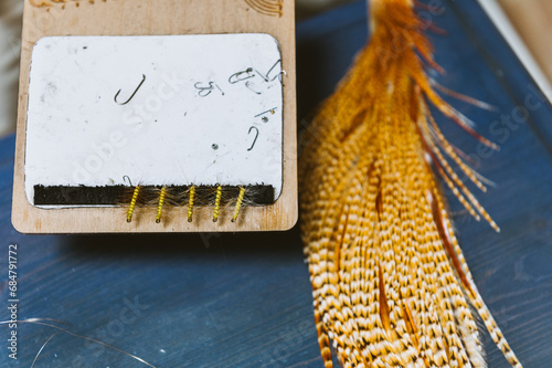 Brown feathers on a blue table used for tying fishing flies, next to a row of tied flies and hooks photo