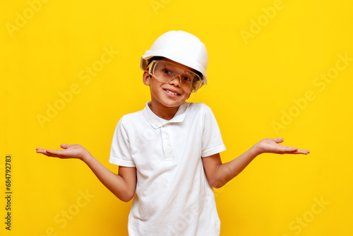 unsure african american boy builder repairman in hard hat and safety glasses shrugs his shoulders on blue background photo