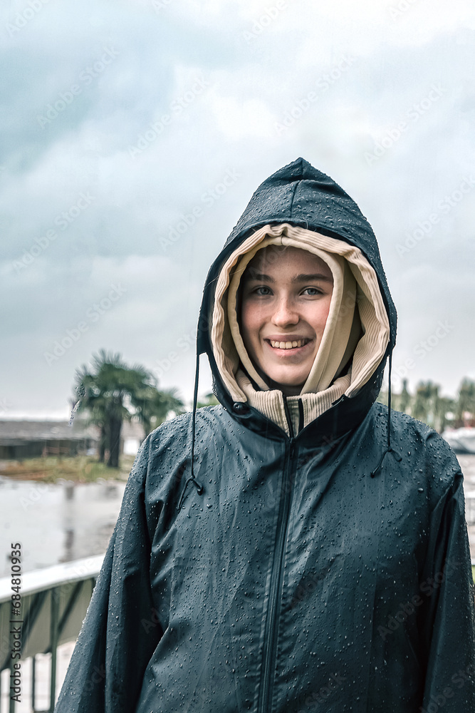 Portrait of a smiling young woman in a raincoat during a storm, hurricane, downpour on the island against the backdrop of the sea Portrait of a smiling young woman in a raincoat during a storm, hurricane, downpour on the island against the backdrop of the sea