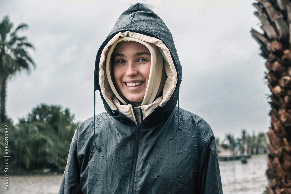 Portrait of a smiling young woman in a raincoat during a storm, hurricane, downpour on the island against the backdrop of the sea Portrait of a smiling young woman in a raincoat during a storm, hurricane, downpour on the island against the backdrop of the sea