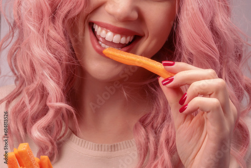 Carrots cut into strips in woman hand. Woman with pink curly hair is eating carrot.