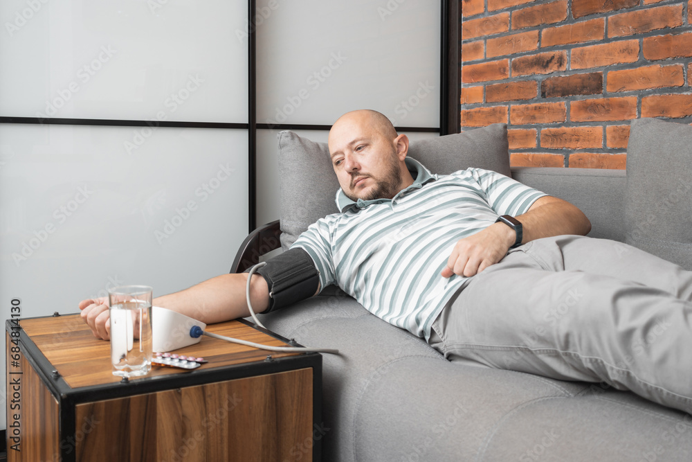 Fototapeta premium Young adult man lying on a sofa at home measures blood pressure on a hand with modern digital tonometer