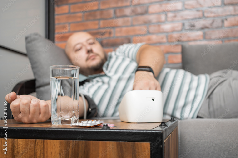 Defocused background with man who has hypertension and headache, lying on sofa at home, measures blood pressure on hand with modern digital tonometer, focus on blister pack with pills on foreground Defocused background with man who has hypertension and headache, lying on sofa at home, measures blood pressure on hand with modern digital tonometer, focus on blister pack with pills on foreground